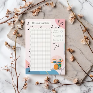 Op de afbeelding: Een roze en wit "Drums tracker" notitieblok met een raster voor het bijhouden van oefensessies. Het notitieblok bevat muzieknoten, een drummer-illustratie en selectievakjes voor oefenniveaus. Decoratieve bloemtakken omlijsten het notitieblok.