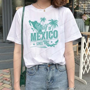 May include: White t-shirt with a teal graphic design featuring the words "Gulf of Mexico" and "Since 1672". The design includes palm trees and a map outline. The shirt is paired with blue denim shorts.
