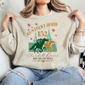 Może przedstawiać: Beżowa bluza z okrągłym dekoltem z grafiką przedstawiającą dwa konie wyścigowe, dżokejów i tekst "Kentucky Derby 152 Churchill Downs Run for the Roses May 3 2026" w kolorze zielonym, złotym i czerwonym. Projekt zawiera ilustracje róż.