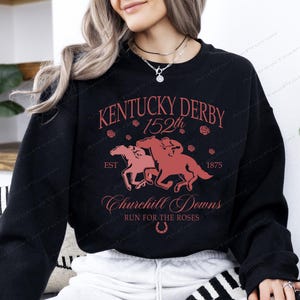 May include: Black sweatshirt with a red graphic featuring the text "Kentucky Derby 152th" and "Churchill Downs". The design includes two horses and riders, roses, and a horseshoe. Est. 1875.