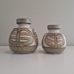Op de afbeelding: Twee keramische vazen met een bolle vorm en een smalle hals. De vazen zijn versierd met een bruin en wit patroon. De grotere vaas heeft een cirkelvormig ontwerp, terwijl de kleinere vaas een bladachtig ontwerp heeft.