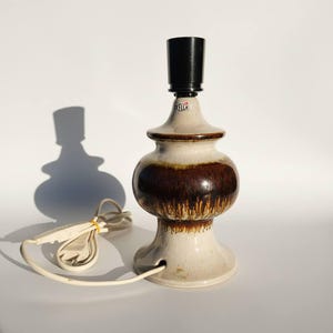 Op de afbeelding: Een keramische tafellamp met een beige en bruin glazuur. De lamp heeft een lamphouder en een netsnoer. Het ontwerp van de lamp is klassiek, vintage. De lamp staat op een wit oppervlak.
