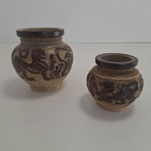 Op de afbeelding: Twee kleine, decoratieve keramische vazen. De vazen zijn bruin met donkerbruine randen en hebben reliëfontwerpen van mythische wezens. De grotere vaas heeft een leeuwachtig figuur, terwijl de kleinere vaas een draakachtig ontwerp heeft.
