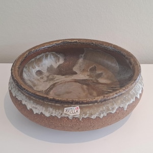 Op de afbeelding: Een ronde keramische kom met een brede rand. De kom heeft een gespikkelde, roodbruine buitenkant en een lichtere binnenkant met witte en bruine marmering. De rand heeft een witte, getextureerde rand. De kom heeft een "Axella"-sticker.