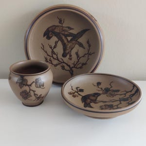 May include: A trio of ceramic tableware in tan with brown bird and floral designs. The set includes a bowl, a small dish, and a cup. The bowl has a dark brown stripe around the rim.