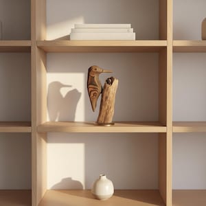 Op de afbeelding: Een houten sculptuur van een vogel met een lange snavel, geplaatst op een stuk drijfhout. De vogel is bruin met een patroon. De sculptuur staat op een goudkleurige basis. Een kleine witte vaas en een stapel boeken zijn ook zichtbaar.