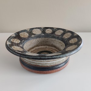 Op de afbeelding: Een handgemaakte keramische kom met een brede, licht uitlopende rand. De rand is versierd met witte stippen op een donkerblauwe achtergrond. De basis van de kom heeft horizontale banden in bruin, zwart en rood. Dit decoratieve stuk is geschikt voor diverse doeleinden.