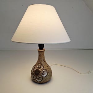 Op de afbeelding: Een keramische tafellamp met een witte lampenkap. De lampvoet is een gespikkelde bruine kleur met een bloemenontwerp. De lamp heeft een zwarte hals en een wit snoer.