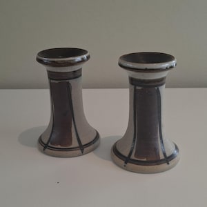 Op de afbeelding: Twee keramische kandelaars. De kandelaars zijn beige met bruine en zwarte accenten. De boven- en onderkant zijn breder dan het middengedeelte. De kandelaars staan op een wit oppervlak.