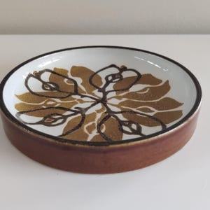 Può includere: Un piatto rotondo in ceramica con base marrone e interno bianco. L'interno presenta un motivo floreale nei toni dell'oro e del marrone. Il bordo è bordato di nero.