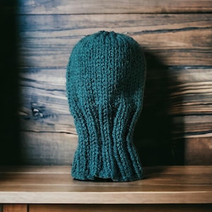 Peut inclure: Une cagoule bleu sarcelle foncé fabriquée en grosse laine tricotée. Le couvre-chef présente une texture côtelée verticale sur toute sa surface, s'étendant du sommet arrondi jusqu'à la zone du cou, offrant un ajustement serré et uniforme.