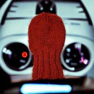 May include: A rust-coloured knitted beanie with a ribbed texture and a rounded crown. The hat features a vertical knit pattern throughout the body and a slightly tighter ribbed cuff at the base, crafted from a soft, fuzzy yarn material.