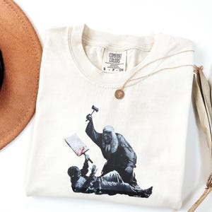 May include: Cream-colored Comfort Colors t-shirt featuring a black and white graphic of a judge with a hammer and a person on the ground. A gold necklace with a pendant and a brown hat are also visible.