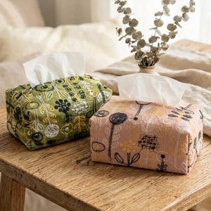 Puede incluir: Dos cubiertas decorativas para cajas de pañuelos con bordados florales. Una es verde oliva con bordados blancos y negros, la otra es rosa rubor con bordados negros y amarillos. Ambas tienen pañuelos blancos y están sobre una superficie de madera.
