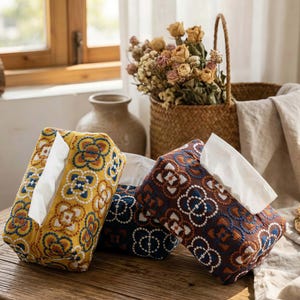 May include: Three decorative tissue box covers with embroidered floral patterns. One is yellow with blue and white accents, another is navy blue with white accents, and the third is brown with blue and white accents. Each box holds white tissues.