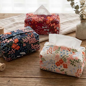 May include: Three decorative tissue box covers with floral patterns. One is red with orange and white flowers, another is navy blue with orange and pink flowers, and the third is cream-colored with embroidered flowers in various colors. White tissues are visible.