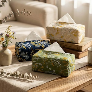 Peut inclure: Trois boîtes à mouchoirs décoratives aux motifs floraux. Une bleue marine, une vert olive et une beige. Chaque boîte présente un motif floral avec des accents jaunes et blancs. Des mouchoirs blancs sont visibles.