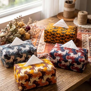 Op de afbeelding: Vier decoratieve tissueboxhoezen met een paardenpatroon in verschillende kleuren: geel, blauw, rood en bruin. Elke doos heeft een wit tissue dat eruit steekt. De dozen staan op een houten tafel.