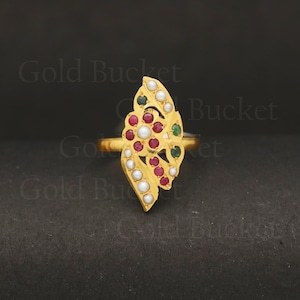 May include: A gold ring featuring a leaf design, embellished with small, round white, red, and green gemstones. The ring is set against a dark background.