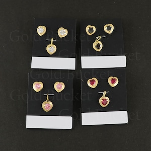 Puede incluir: Cuatro conjuntos de joyas en forma de corazón sobre tarjetas negras, cada uno con dos pendientes y un colgante. Los conjuntos presentan piedras transparentes, negras, rosa claro y rojo oscuro engastadas en metal dorado con borde texturizado.