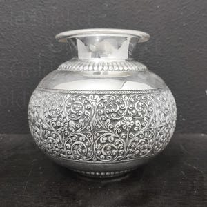 May include: A silver-colored, round vase with intricate floral and scrollwork patterns. The vase has a wide, decorative band near the top and a flared rim. The detailed design covers the majority of the vase's surface, creating a textured appearance.