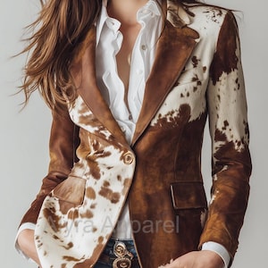 May include: A brown and white cowhide blazer with a two-button closure and two front pockets. The blazer is worn over a white button-down shirt and blue jeans. The brand name "Alyra Apparel" is visible on the blazer.