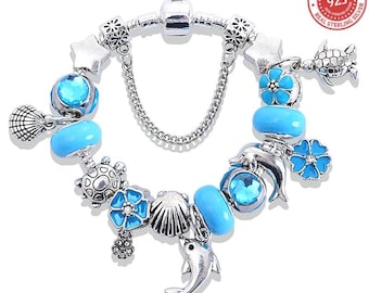 May include: A silver charm bracelet with various ocean-themed charms, including dolphins, turtles, seashells, and blue beads. The bracelet has a chain and a clasp. The charms are detailed and the bracelet is set against a white background.