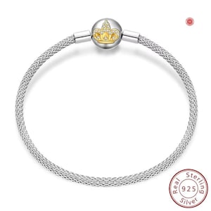 May include: A silver bracelet with a woven chain design. The bracelet features a gold-coloured charm with a crown design, adorned with small, clear gemstones. The charm is centered on the bracelet. The bracelet is made of 925 sterling silver.