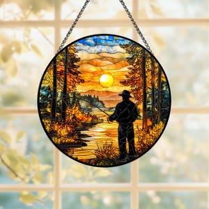 Puede incluir: Panel redondo de vidrieras que representa a un pescador lanzando su línea al atardecer. La escena presenta árboles, montañas y un cielo vibrante naranja y amarillo. El panel está enmarcado en negro y cuelga de una cadena.