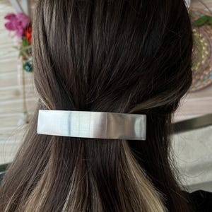 Könnte beinhalten: Eine rechteckige Haarspange in Weiß-, Grau- und Blassblautönen ist in dunkelbraunes Haar geklemmt. Die Spange hat eine glatte, polierte Oberfläche und ist horizontal am Hinterkopf positioniert.