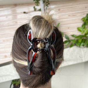 Peut inclure: Une pince à cheveux en écaille de tortue marron foncé et rouge, fixant un chignon. La pince a une charnière en métal doré et un mécanisme à ressort. Cet accessoire pour cheveux est une façon élégante de relever les cheveux.