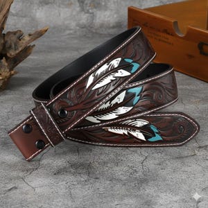 May include: A brown leather belt with a carved feather design. The feathers are white and turquoise, with white stitching along the edges. The belt has a dark brown interior and a silver buckle.