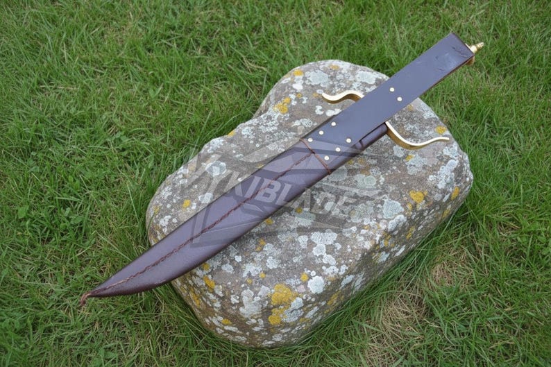 Puede incluir: Una funda de cuero marr&oacute;n oscuro con detalles dorados y un mango a juego. La funda descansa sobre una gran roca cubierta de l&iacute;quenes, con hierba verde en el fondo. Es una espada o un cuchillo.