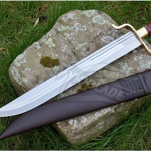 Puede incluir: Una espada de hoja plateada con empu&ntilde;adura marr&oacute;n y dorada, apoyada sobre una piedra. La espada tiene una guarda curva y va acompa&ntilde;ada de una vaina de cuero marr&oacute;n. La espada se exhibe sobre un lecho de hierba verde.