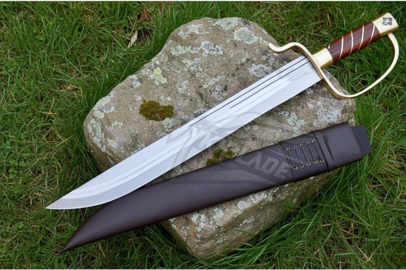 Puede incluir: Una espada de hoja plateada con empu&ntilde;adura marr&oacute;n y dorada, apoyada sobre una piedra. La espada tiene una guarda curva y va acompa&ntilde;ada de una vaina de cuero marr&oacute;n. La espada se exhibe sobre un lecho de hierba verde.
