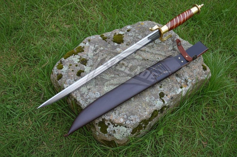 Puede incluir: Una espada decorativa con un mango marr&oacute;n y dorado, apoyada sobre una piedra. La hoja es plateada y la vaina es negra. La espada se exhibe sobre una piedra con hierba verde en el fondo.