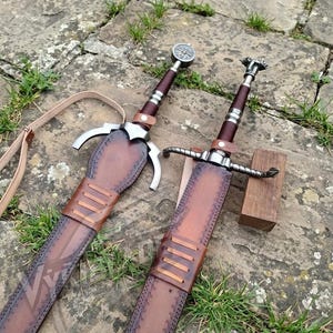 May include: Two swords with dark brown leather-wrapped grips and metallic pommels, each housed in a matching brown leather scabbard featuring decorative stitching, leather strap accents, and a light-coloured leather carrying belt attachment.