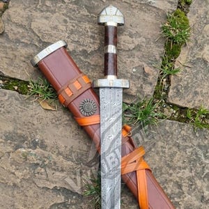 May include: A sword with a patterned Damascus steel blade, a dark wood handle, and silver-toned metal fittings. The sword is accompanied by a brown leather scabbard featuring orange leather straps and a circular metal decorative emblem.