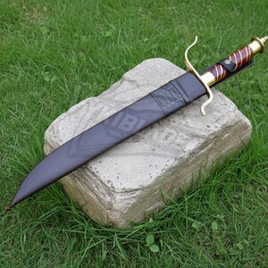 Puede incluir: Un cuchillo grande y oscuro con una hoja curva y un mango decorativo, descansando en una funda negra. El mango presenta detalles dorados y marrones. El cuchillo est&aacute; colocado sobre una piedra, con hierba verde en el fondo.