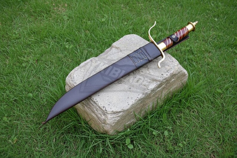 Puede incluir: Un cuchillo grande y oscuro con una hoja curva y un mango decorativo, descansando en una funda negra. El mango presenta detalles dorados y marrones. El cuchillo est&aacute; colocado sobre una piedra, con hierba verde en el fondo.