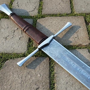May include: A silver and brown sword with a leather-wrapped handle and a detailed blade. The blade has an etched design and text. The sword is lying on a stone surface with grass.