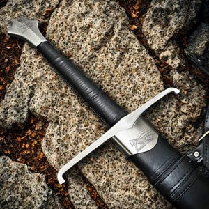 Puede incluir: Primer plano de una espada con hoja y guarda plateadas, y un mango envuelto en cuero negro. La espada descansa sobre una superficie de piedra texturizada. La palabra "Vykblade" es visible en la guarda.