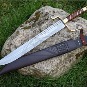 Puede incluir: Una espada de hoja plateada con un mango marr&oacute;n y dorado descansa sobre una gran roca, junto a una vaina de cuero marr&oacute;n oscuro. La espada tiene una guarda curva y un patr&oacute;n decorativo en la hoja. La vaina est&aacute; detallada con costuras y una correa de cuero.