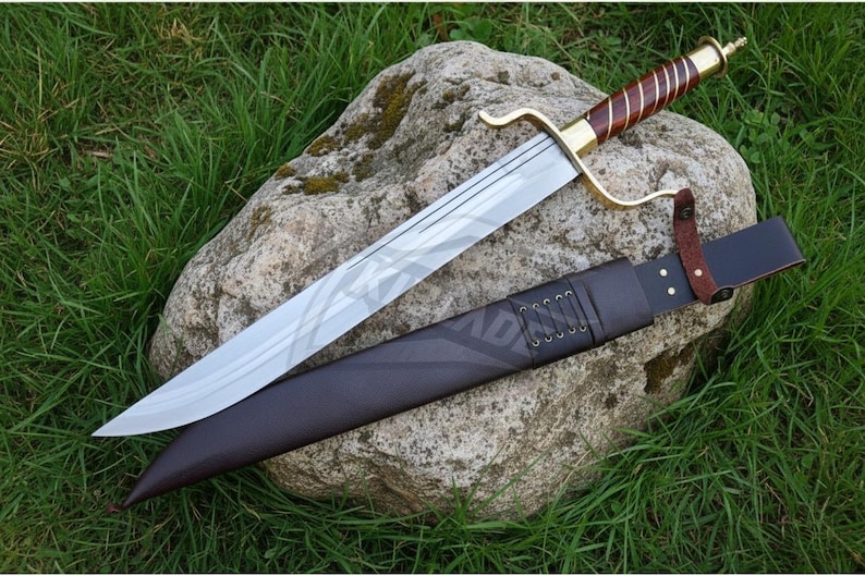 Puede incluir: Una espada de hoja plateada con un mango marr&oacute;n y dorado descansa sobre una gran roca, junto a una vaina de cuero marr&oacute;n oscuro. La espada tiene una guarda curva y un patr&oacute;n decorativo en la hoja. La vaina est&aacute; detallada con costuras y una correa de cuero.