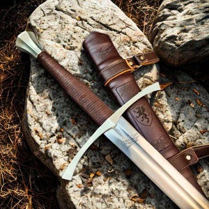 Puede incluir: Una espada medieval con una hoja plateada y un mango envuelto en cuero marrón descansa sobre una superficie de piedra. La espada va acompañada de una funda de cuero marrón con hebilla y diseño en relieve. La hoja tiene el texto "Vykiblade".