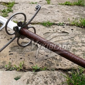 May include: A long, silver sword with a brown, textured handle and ornate metal crossguard. The sword is lying on a stone surface with grass and small plants around it. The blade is sharp and the handle is wrapped in a woven pattern.