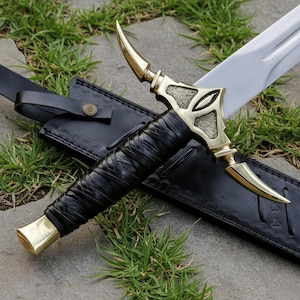May include: A detailed image of a decorative sword with a gold-colored hilt and a black leather-wrapped handle. The sword's blade is silver, and it rests in a black leather sheath. The hilt features intricate designs, and the handle is tightly wrapped. The sword is displayed outdoors.