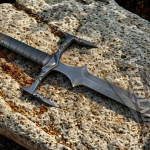 May include: A detailed, silver-colored sword with a textured handle and ornate crossguard rests on a rough, gray stone. The blade has a pointed tip and decorative etching. The sword is a fantasy-themed item.
