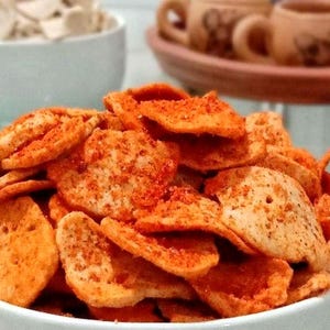 May include: A white bowl overflowing with seasoned, orange-dusted chips. Another bowl in the background holds similar, unseasoned chips. In the background are small brown mugs on a terracotta tray, suggesting a snack or appetizer.