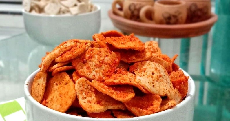 May include: A white bowl overflowing with seasoned, orange-dusted chips. Another bowl in the background holds similar, unseasoned chips. In the background are small brown mugs on a terracotta tray, suggesting a snack or appetizer.